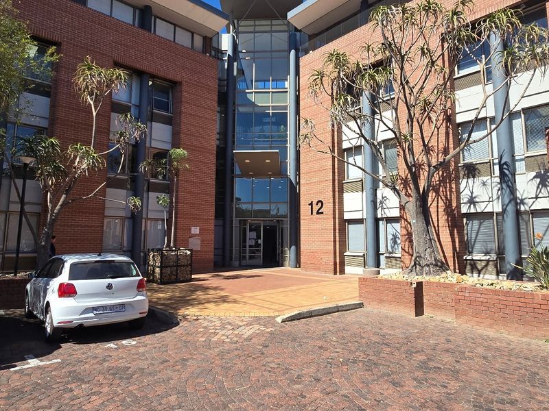 Front elevation of a multi-story commercial office building featuring a brick facade, glass entrance atrium, and paved parking area.