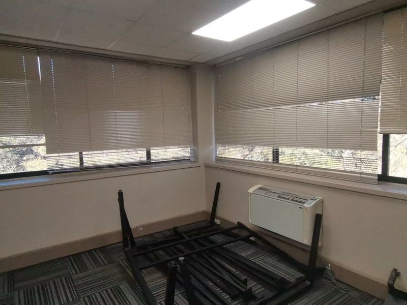 A vacant office space featuring carpet tiles, recessed lighting, wall-mounted HVAC unit, and large windows with venetian blinds.