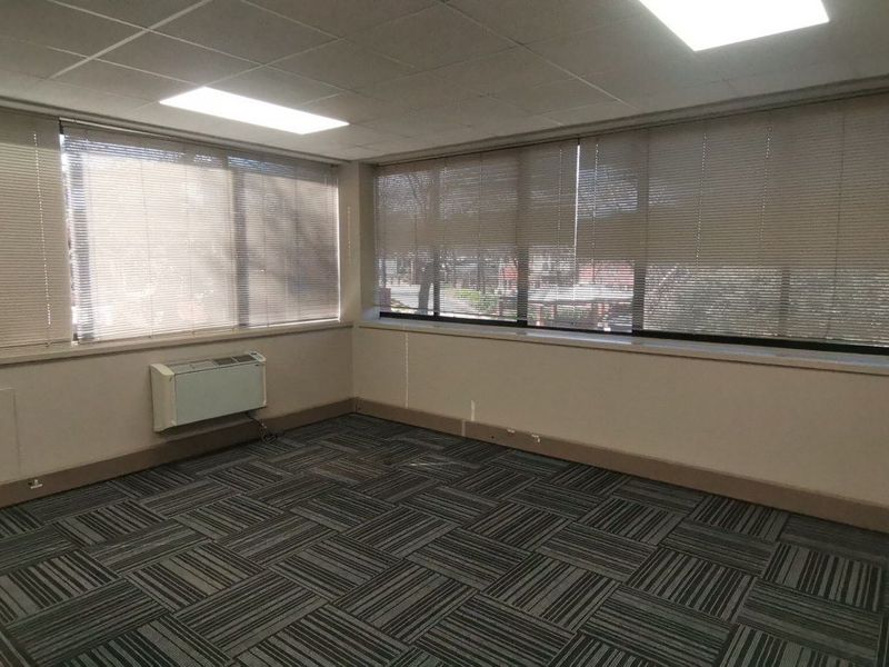 A vacant commercial office space featuring large windows with blinds, carpet tile flooring, a suspended ceiling, and a wall-mounted HVAC unit.