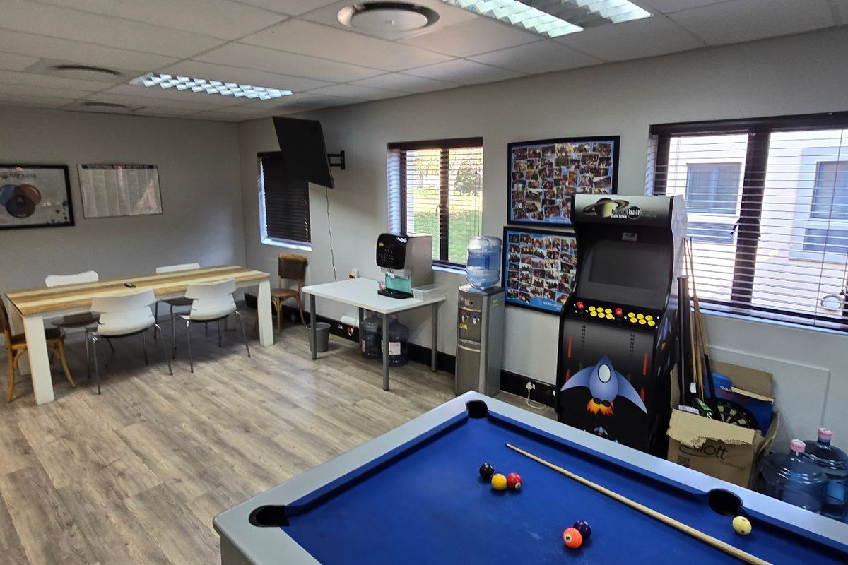 A multi-purpose commercial break room featuring laminate flooring, recessed lighting, and large windows with venetian blinds, currently set up with recreational and dining areas.