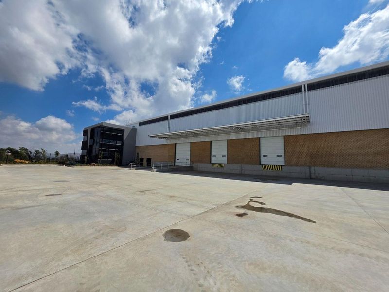 Industrial warehouse exterior featuring multiple loading bays with roller shutter doors, a concrete yard, and a modern multi-level office facade.