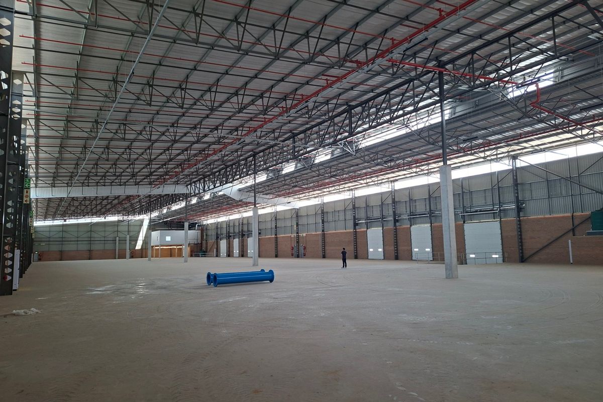 Expansive industrial warehouse space featuring high ceilings with exposed steel trusses, fire sprinkler systems, concrete flooring, and multiple roller shutter doors.