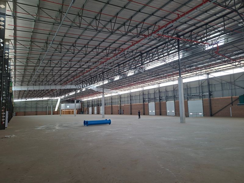 Expansive industrial warehouse space featuring high ceilings with exposed steel trusses, fire sprinkler systems, concrete flooring, and multiple roller shutter doors.
