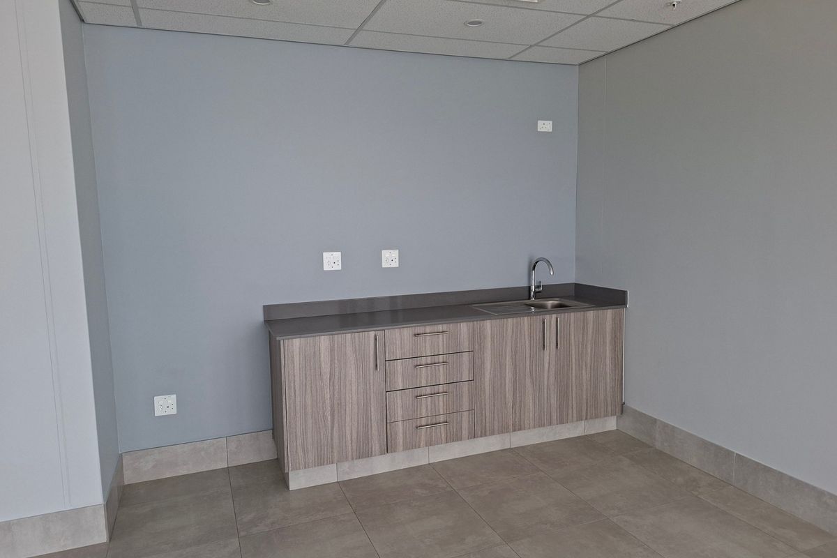 A clean, modern kitchenette area featuring wood-grain cabinetry, a stone countertop with an integrated sink, and neutral tiled flooring in an industrial office space.
