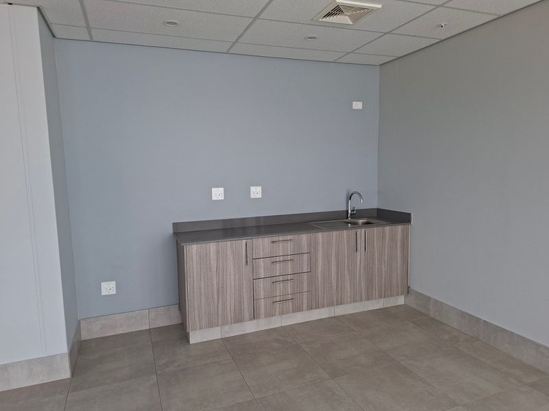 A clean, modern kitchenette area featuring wood-grain cabinetry, a stone countertop with an integrated sink, and neutral tiled flooring in an industrial office space.