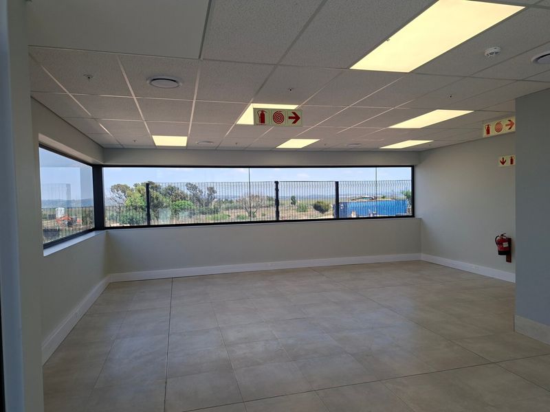 A bright, empty office space featuring large windows with exterior views, modern tiled flooring, and a clean suspended ceiling with recessed lighting.