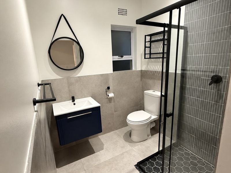A modern, renovated bathroom featuring a wall-mounted vanity, walk-in shower with black frame, and contemporary grey tiling.