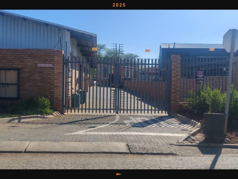 Commercial property entrance featuring a secure metal gate, brick perimeter wall with electric fencing, and a paved driveway leading to the industrial facility.