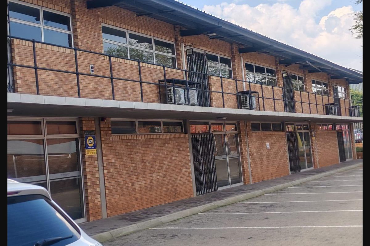 A two-story commercial office building featuring a face brick facade, aluminum window frames, and a balcony with metal railings, situated above a paved parking area.