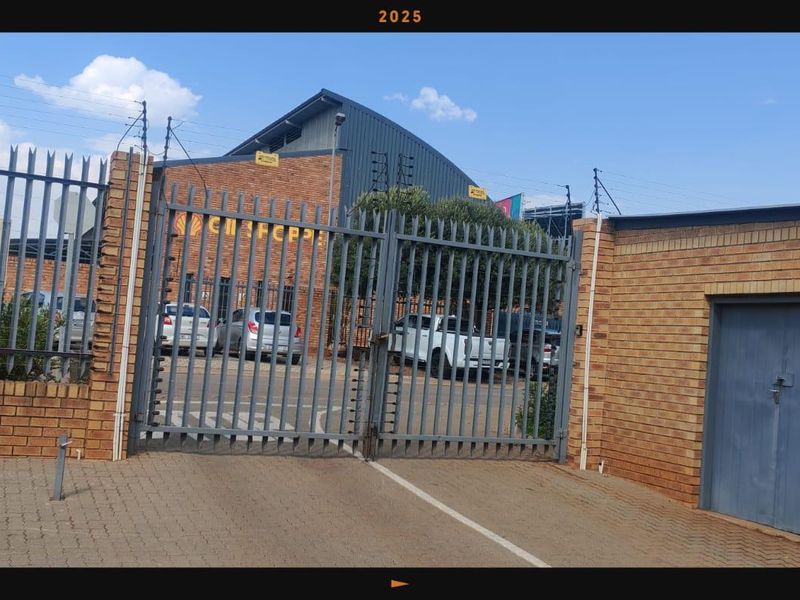 Commercial property entrance featuring a secure steel gate, brick perimeter wall with electric fencing, and paved driveway access to the industrial building.