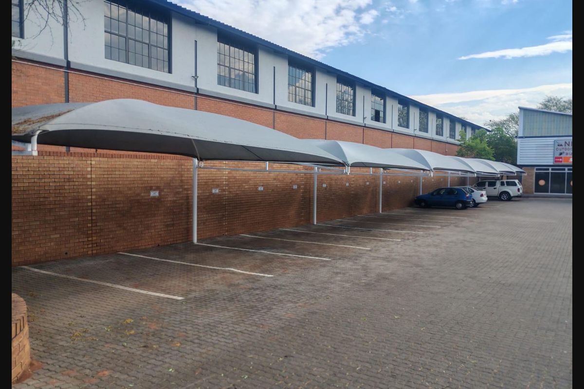 Commercial parking area featuring a row of shade cloth carports, paved surfacing, and a brick boundary wall against an industrial building facade.