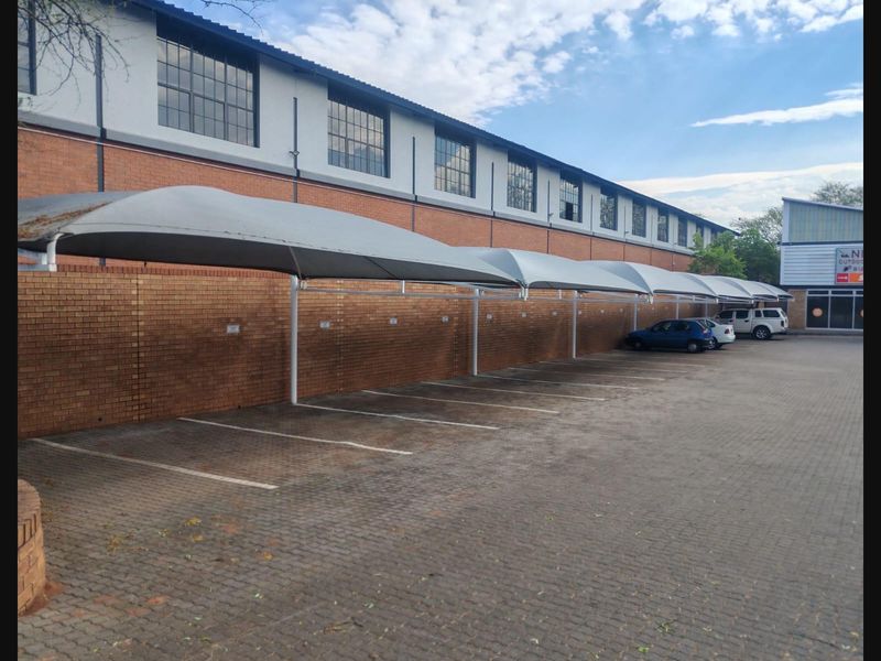 Commercial parking area featuring a row of shade cloth carports, paved surfacing, and a brick boundary wall against an industrial building facade.