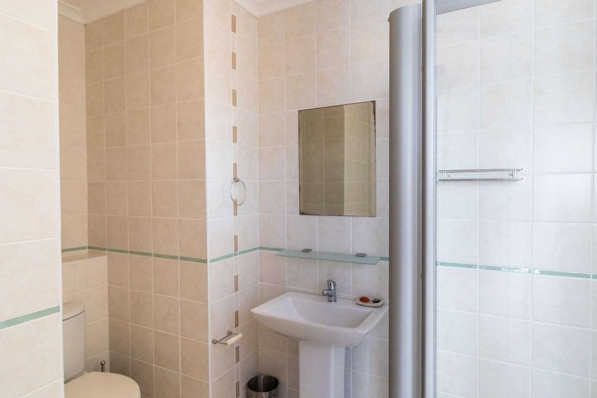 A functional bathroom featuring a pedestal sink, wall-mounted mirror, glass shelving, and a shower enclosure with neutral wall tiling.