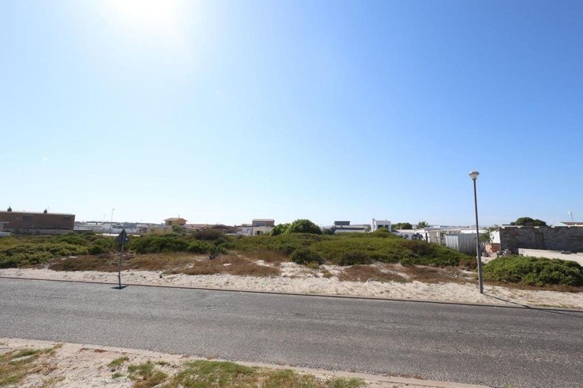 A street-level view of a vacant residential plot featuring sandy terrain, native coastal vegetation, and adjacent neighborhood housing.