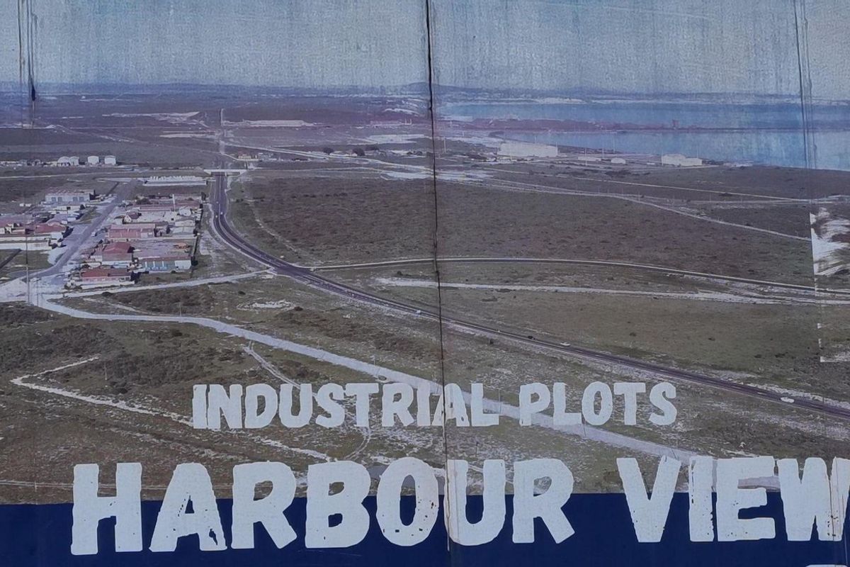 A large outdoor billboard displaying an aerial view of industrial plots and harbour surroundings, showing significant signs of weathering and wear.