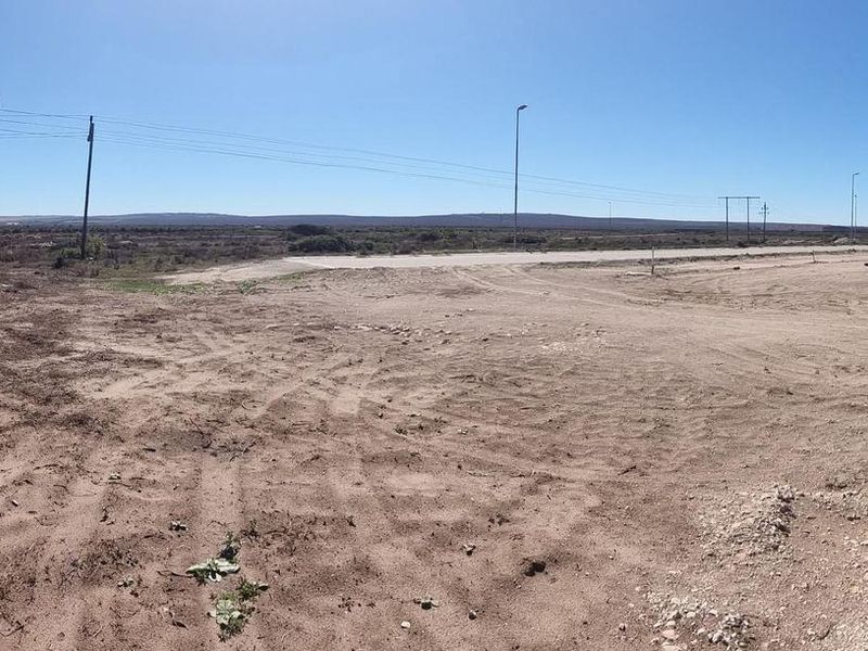 A wide panoramic view of an undeveloped commercial plot featuring dirt terrain, utility poles, and distant horizon lines under a clear sky.