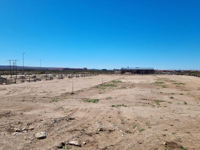 A large, undeveloped commercial plot of land with sandy terrain, marked boundaries, and distant industrial structures under a clear blue sky.