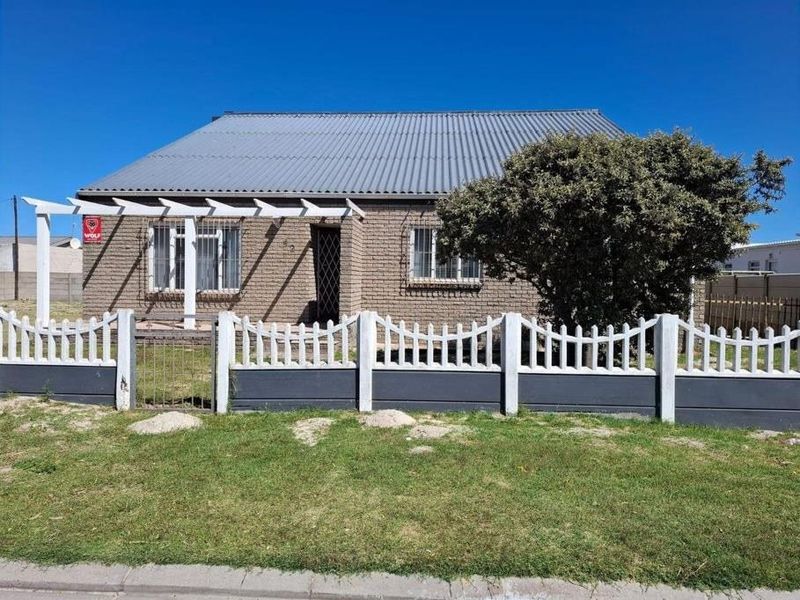 Front view of a single-story brick house featuring a corrugated metal roof, security bars on windows, and a front garden with a precast concrete fence.
