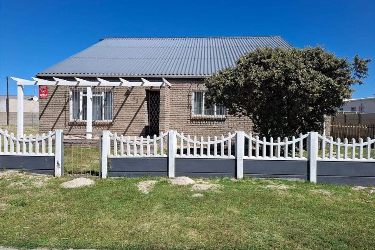 Front view of a single-story brick house featuring a corrugated metal roof, security bars on windows, and a front garden with a precast concrete fence.