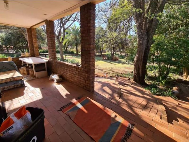 A covered outdoor patio featuring brick pillars and terracotta tile flooring, overlooking a spacious, tree-lined garden area.