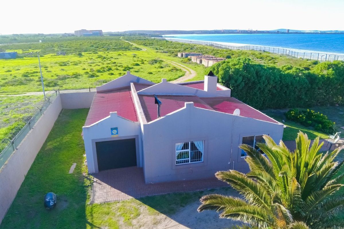 Aerial view of a coastal property featuring a metal roof, attached garage, and surrounding garden area with proximity to the beach.