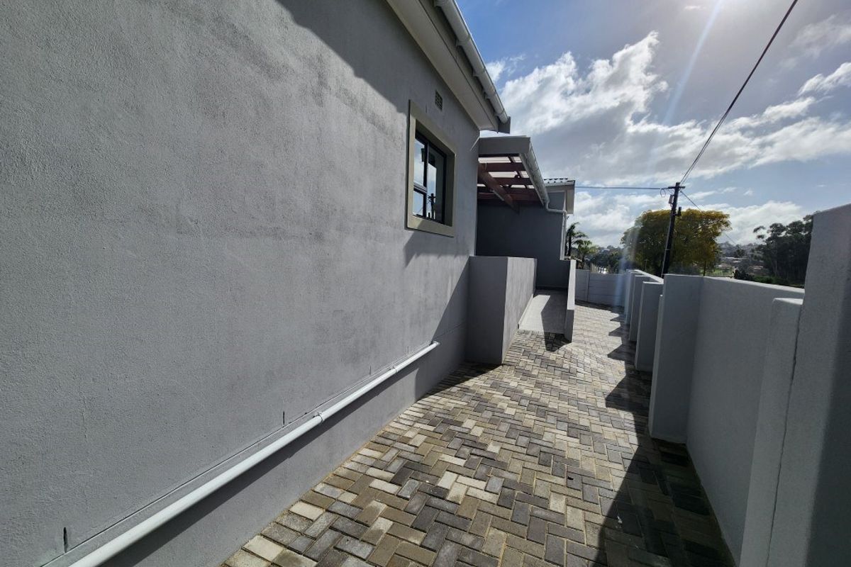 A side exterior walkway featuring paved brick flooring, rendered grey walls, and a covered patio area in the background.