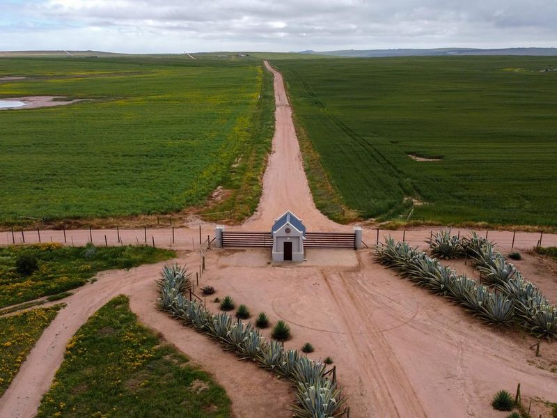 Aerial view of the farm entrance featuring a secure gated access point with a small guardhouse, set against expansive green agricultural fields.