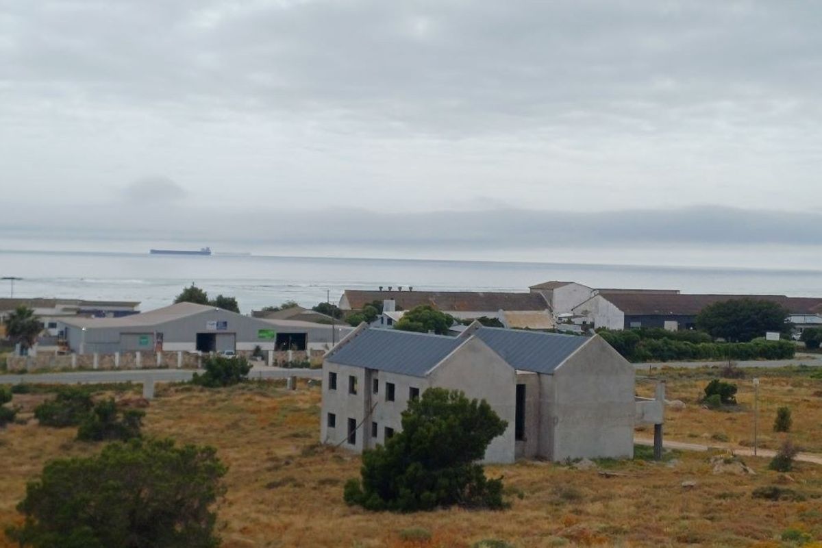 An elevated view of a vacant residential plot featuring an unfinished two-story structure with a metal roof, set against a coastal landscape.