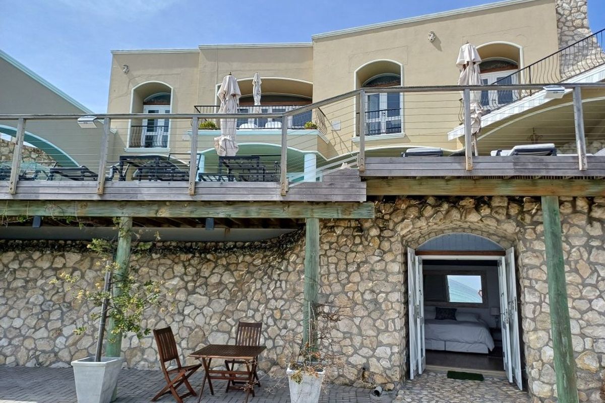 Rear elevation featuring a raised timber deck, stone-clad lower level, and a paved patio area with direct access to a ground-floor bedroom.