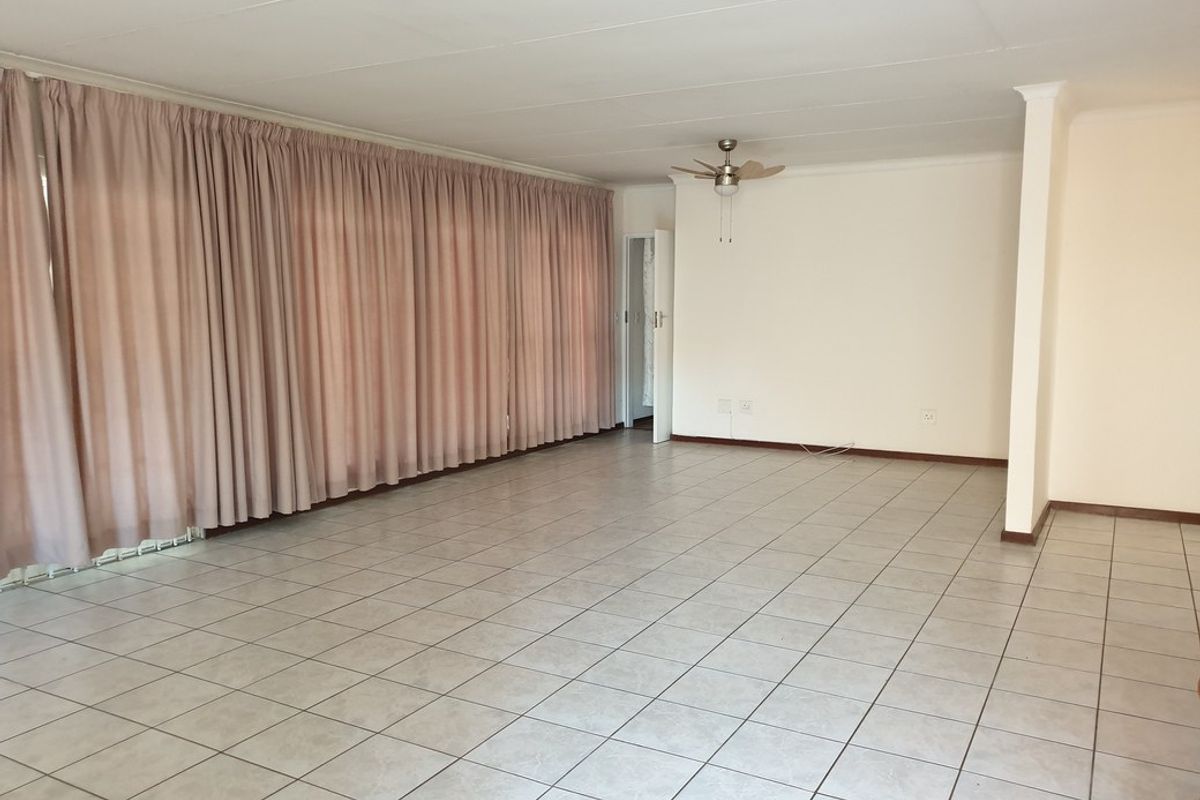 A spacious, open-plan living area featuring neutral tiled flooring, a ceiling fan, and large windows covered by full-length curtains.