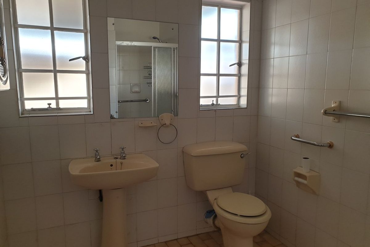A dated bathroom featuring a pedestal basin, toilet, and tiled walls with integrated grab rails and a shower enclosure visible in the mirror reflection.