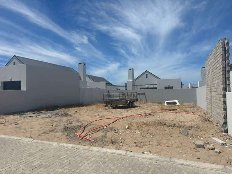 A vacant residential plot featuring a paved street frontage, boundary walls, and surrounding modern housing developments.