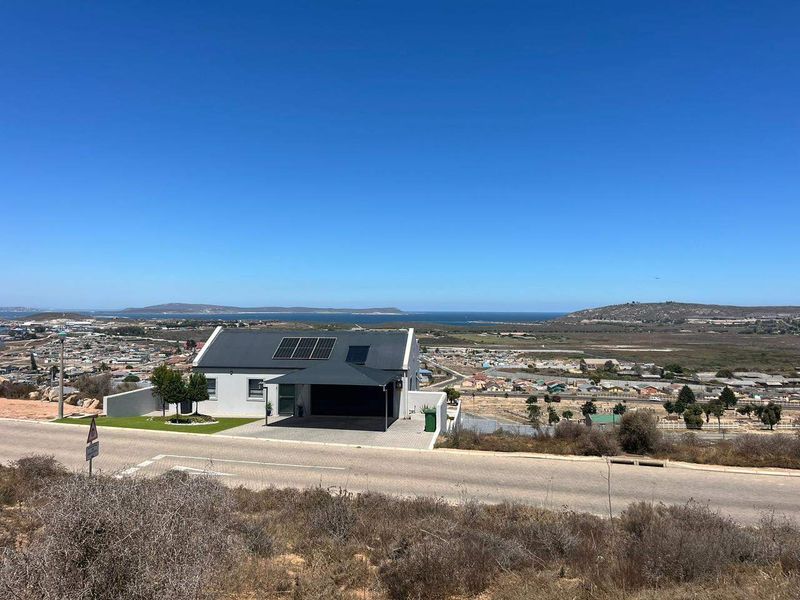 Elevated aerial view of a modern residential property featuring solar panels, a paved driveway, and a covered carport, overlooking a coastal landscape.
