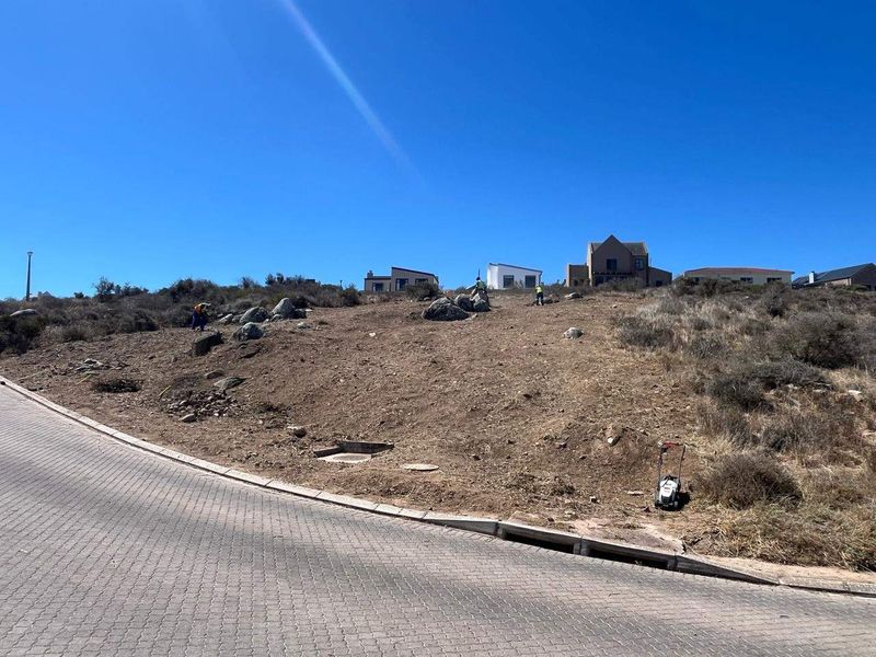 A vacant residential plot featuring natural terrain, rock outcrops, and direct access to a paved street with existing stormwater infrastructure.