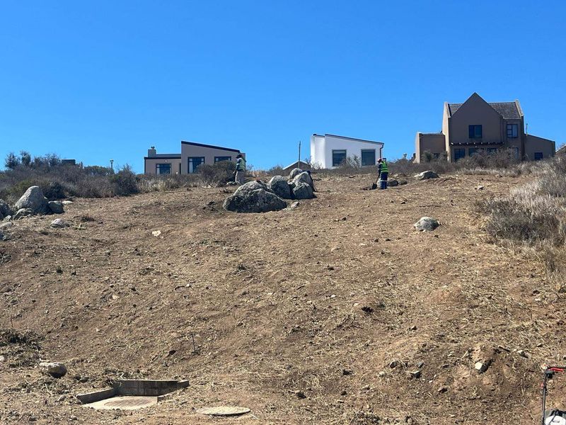 A sloped residential vacant land plot featuring natural rock formations and utility access points, situated in an established neighborhood.