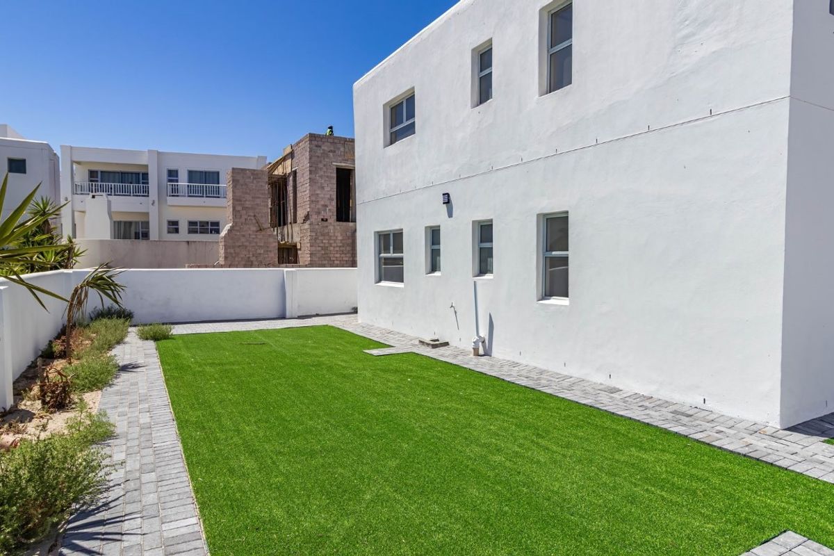 A clean rear exterior view featuring a white rendered facade, paved walkways, and a well-maintained artificial lawn area.