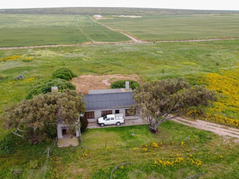 Aerial view of a rural residential property featuring a dwelling with a corrugated metal roof, surrounded by expansive open fields and natural vegetation.