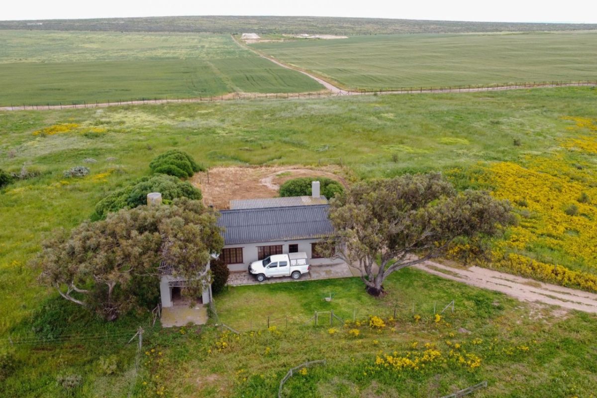 Aerial view of a rural residential property featuring a dwelling with a corrugated metal roof, surrounded by expansive open fields and natural vegetation.