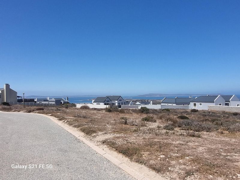 A street-level view of a vacant residential plot in a coastal estate, featuring surrounding neighborhood homes and distant ocean views.