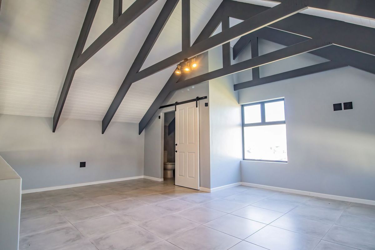 Spacious loft-style bedroom featuring vaulted ceilings with exposed dark beams, tiled flooring, and an en-suite bathroom access via a sliding barn door.