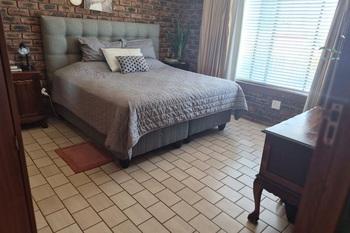 A bedroom featuring an exposed brick feature wall, tiled flooring, and large window with fitted blinds.