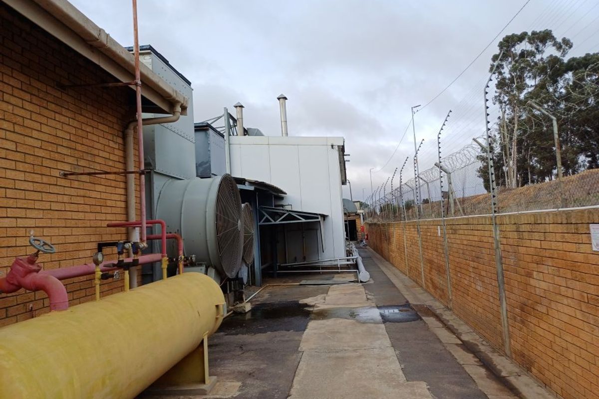 Exterior side passage of an industrial facility featuring large ventilation fans, industrial piping, and a perimeter brick wall with security fencing.