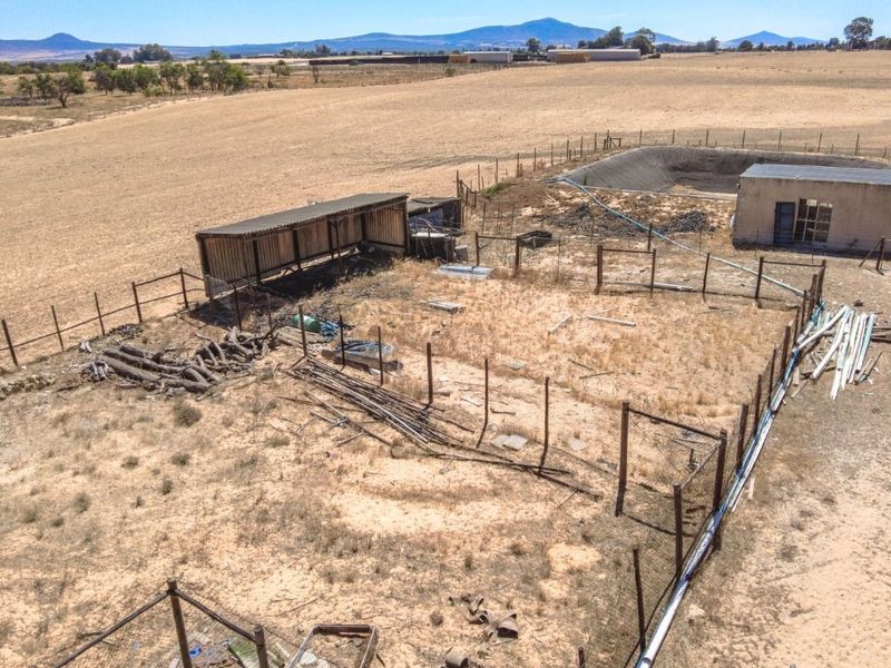 Aerial view of a farm property featuring a livestock shelter, fenced enclosures, an outbuilding, and a water reservoir set within open agricultural land.