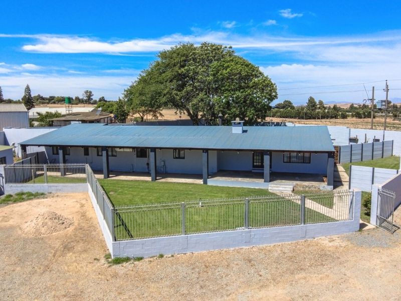 Aerial view of a farm property featuring a single-story dwelling with a covered patio, enclosed by perimeter fencing and concrete boundary walls.