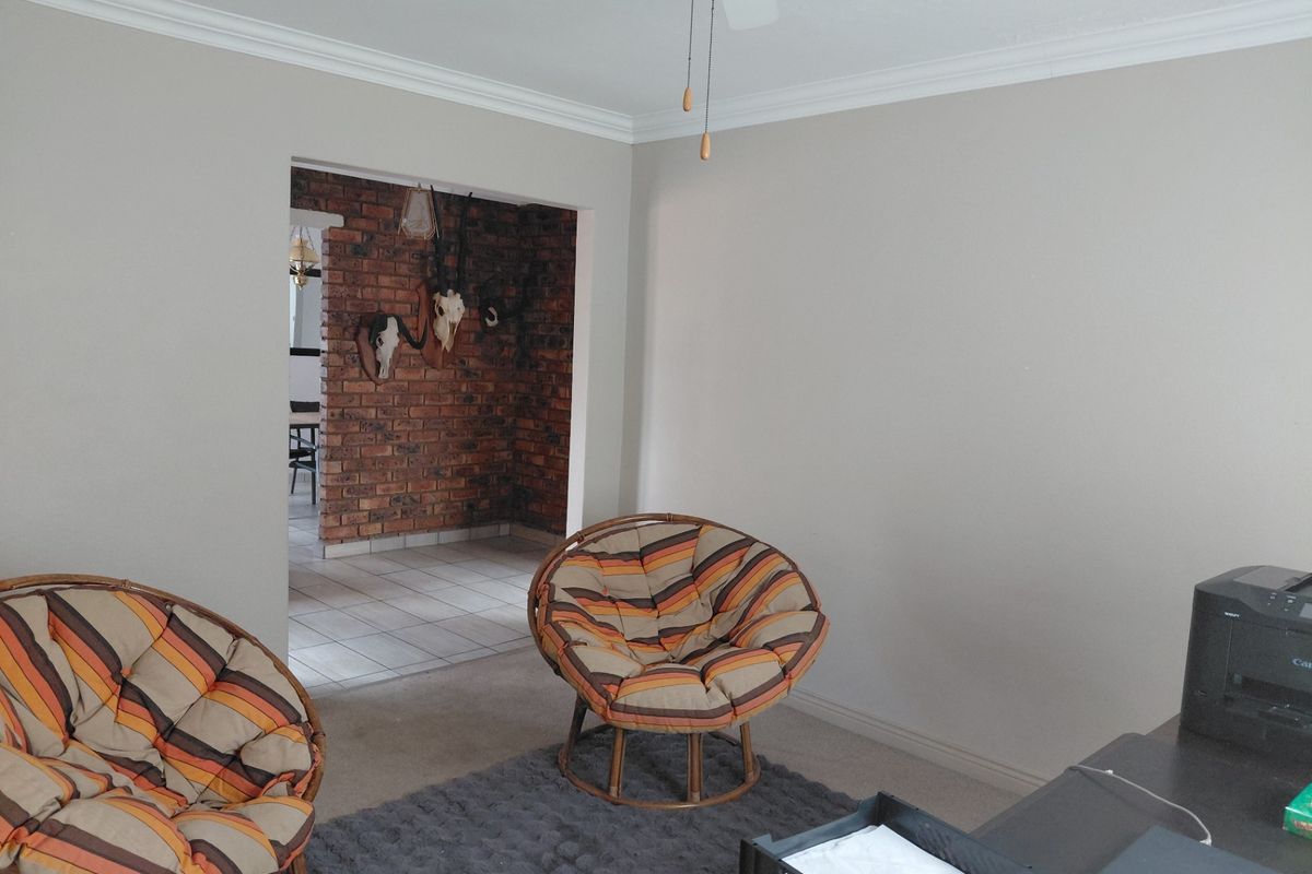 A living room featuring neutral walls, crown molding, and a ceiling fan, with an open transition to an adjacent room with exposed brick wall detailing.