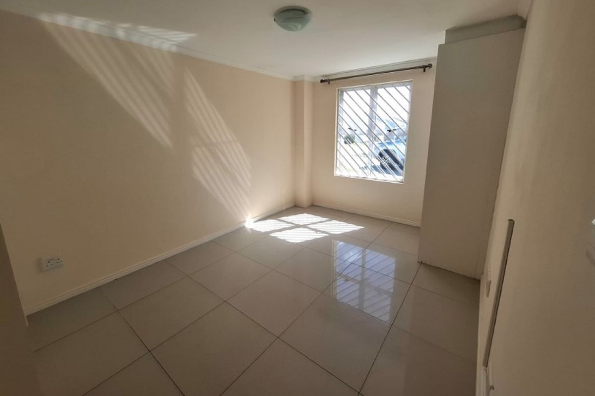 A bright, empty bedroom featuring neutral wall paint, polished floor tiles, and a window fitted with security bars.