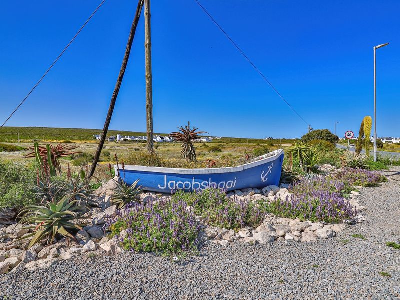 A street-level view of the Jacobsbaai entrance area featuring a decorative boat sign, gravel verge, and local landscaping.