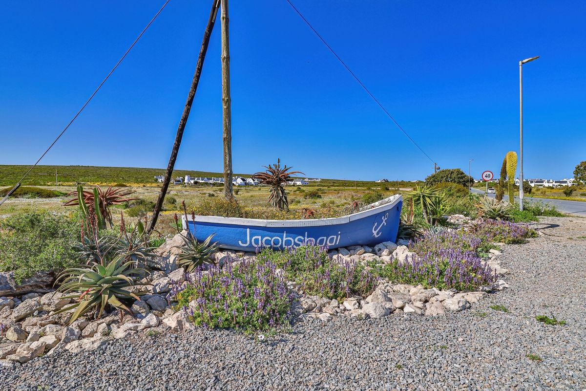 A street-level view of the Jacobsbaai entrance area featuring a decorative boat sign, gravel verge, and local landscaping.