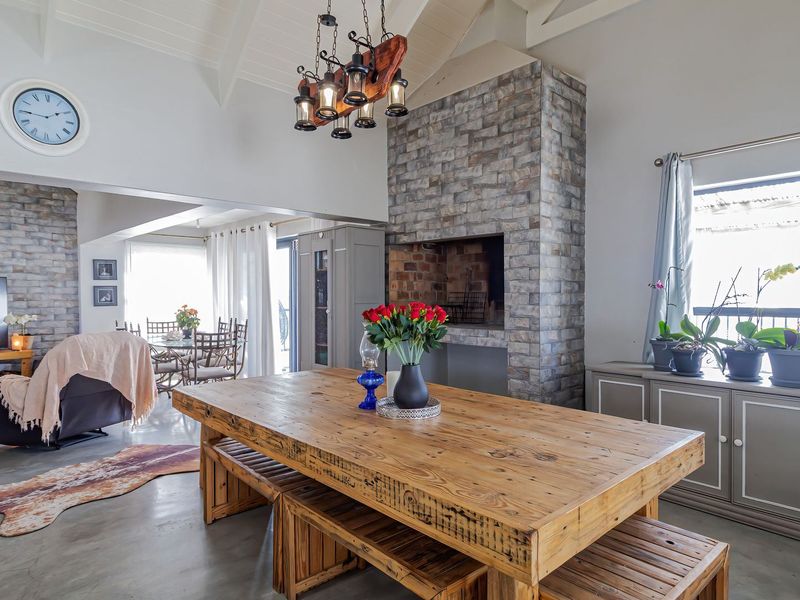 Spacious open-plan living and dining area featuring a vaulted ceiling with exposed beams, polished concrete floors, and a prominent brick fireplace with indoor braai.