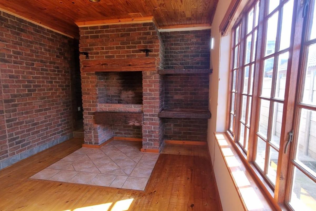 A rustic living room featuring a prominent brick fireplace, timber-clad ceiling, and large wooden-framed windows, offering a warm and character-filled space.